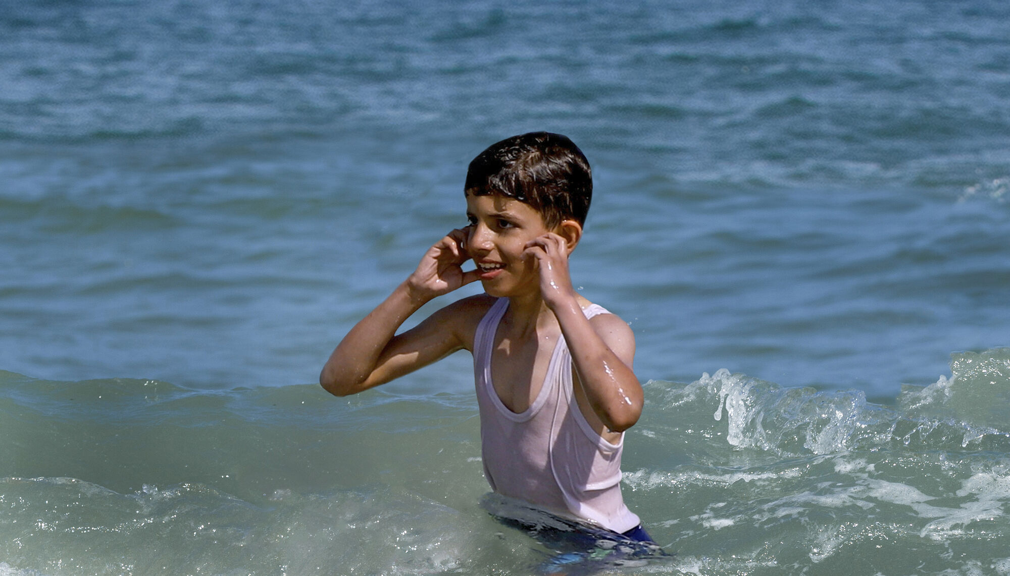 A boy stands waist-deep in the sea off Gaza, cupping his hands to his ears and smiling, as a wave breaks around him.
